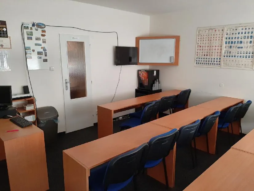 Az autósiskola tanterme: kék székes padok, televízió, fehér tábla és közlekedési táblákkal teli tábla a falon